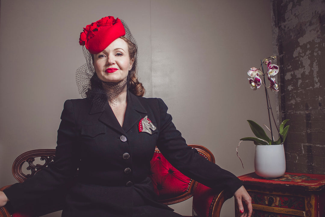 lady wearing a red hat, a black vintage suit, sitting on a sofa next to an orchid