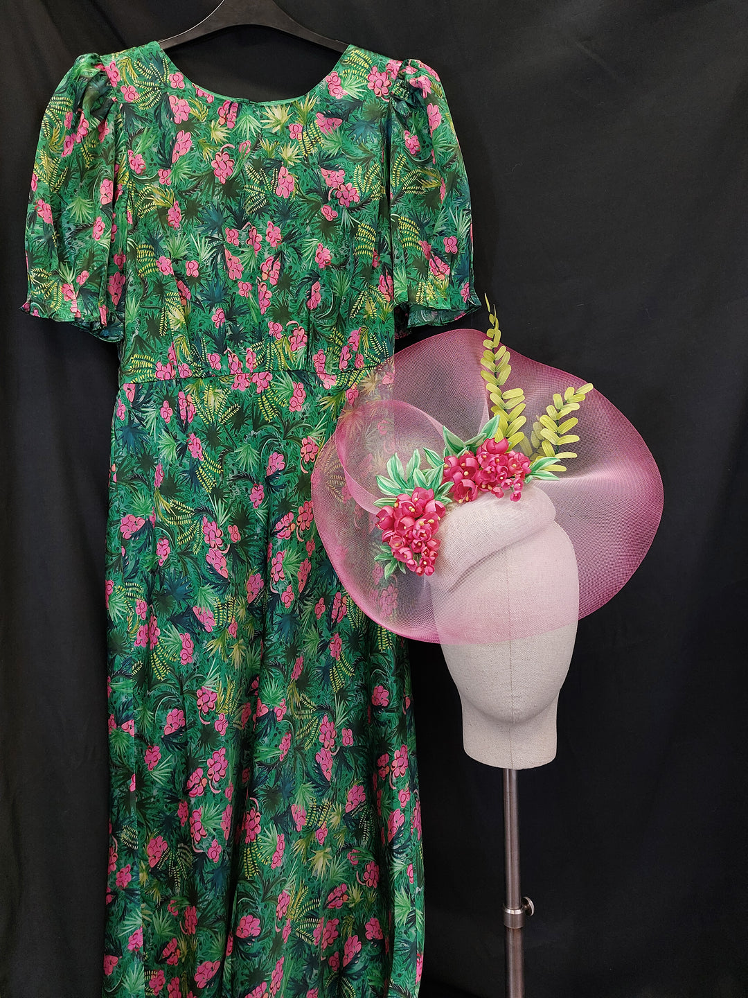 Green floral-patterned dress with a pink flower hat in foreground against a black background