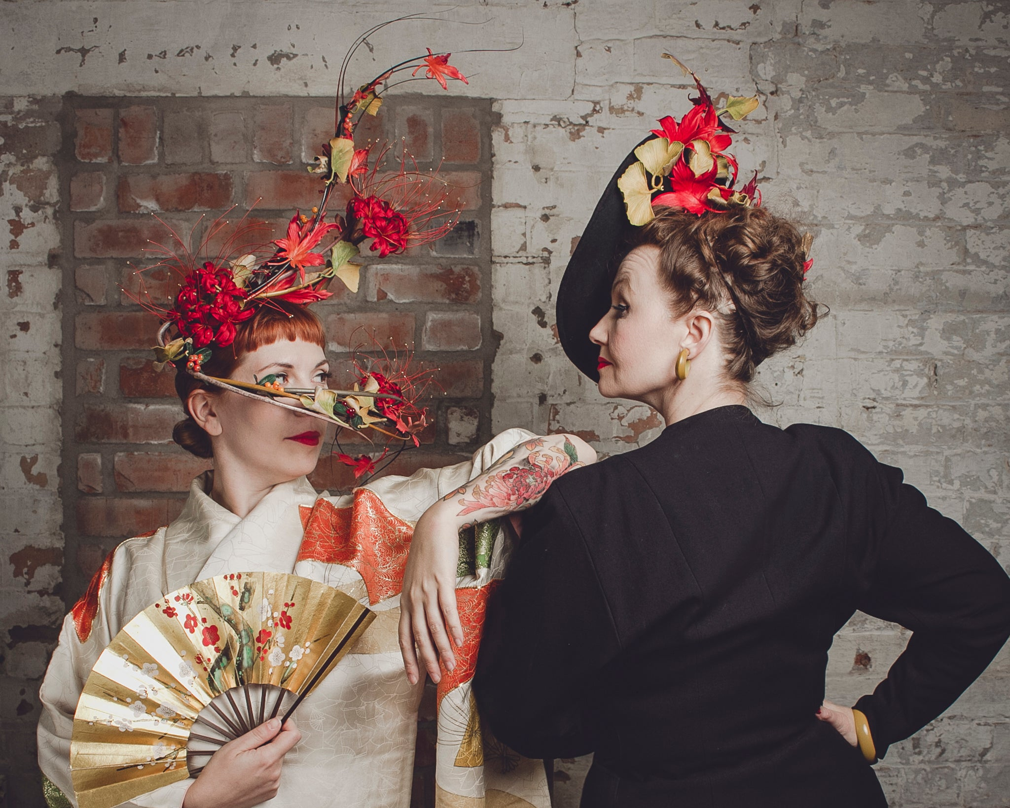 Two women in elaborate hats with floral decorations against a brick wall.