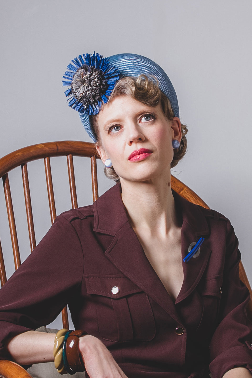 Woman wearing a maroon outfit with a blue hat featuring decorative elements, sitting on a wooden chair.