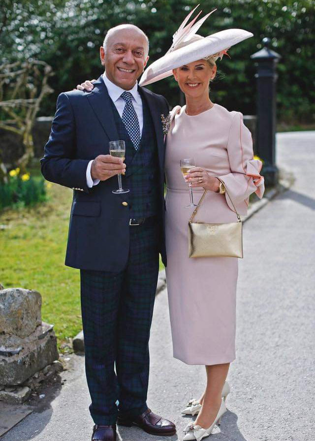 Man and woman posing together outdoors, both holding drinks, with a garden background.