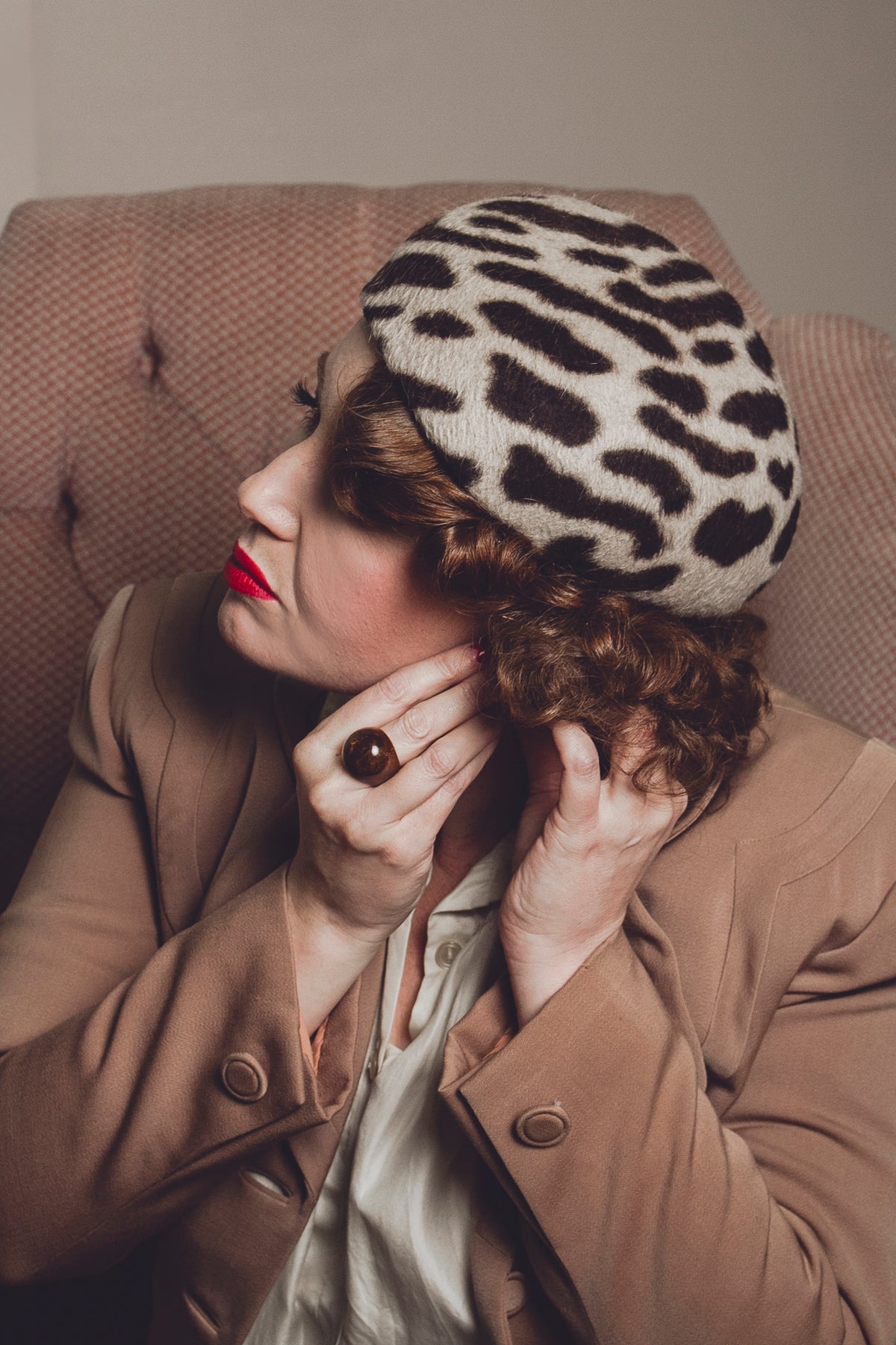 A woman wearing a beige and black leopard-print knit beret, styled with a brown blazer and a light-colored blouse, sitting in a tufted armchair.