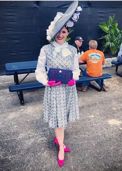 Person in a light blue dress with a large hat, standing outdoors with people and a bench in the background.