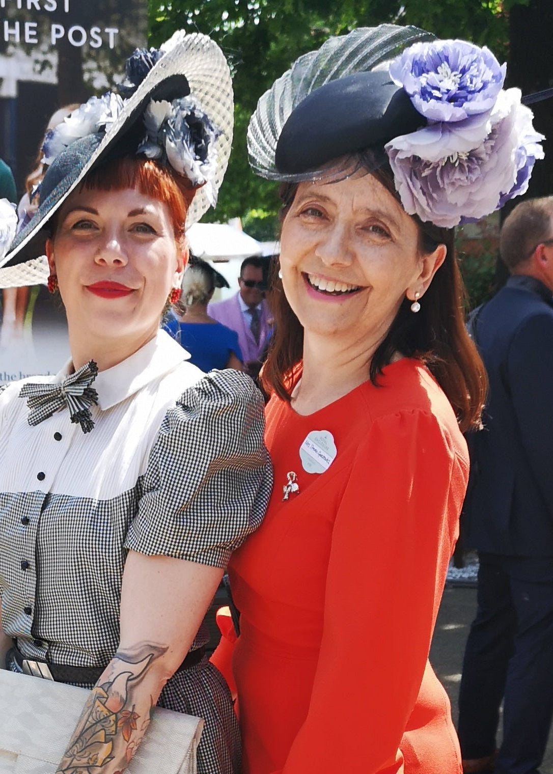 Two women wearing large floral hats, one in a red dress and the other in a checked outfit, standing outdoors.