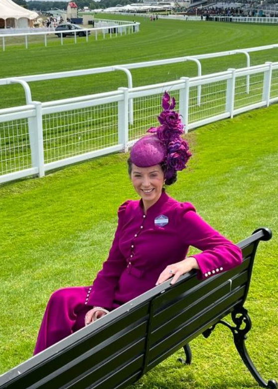 Woman in a purple outfit and hat sitting on a bench at a racecourse.