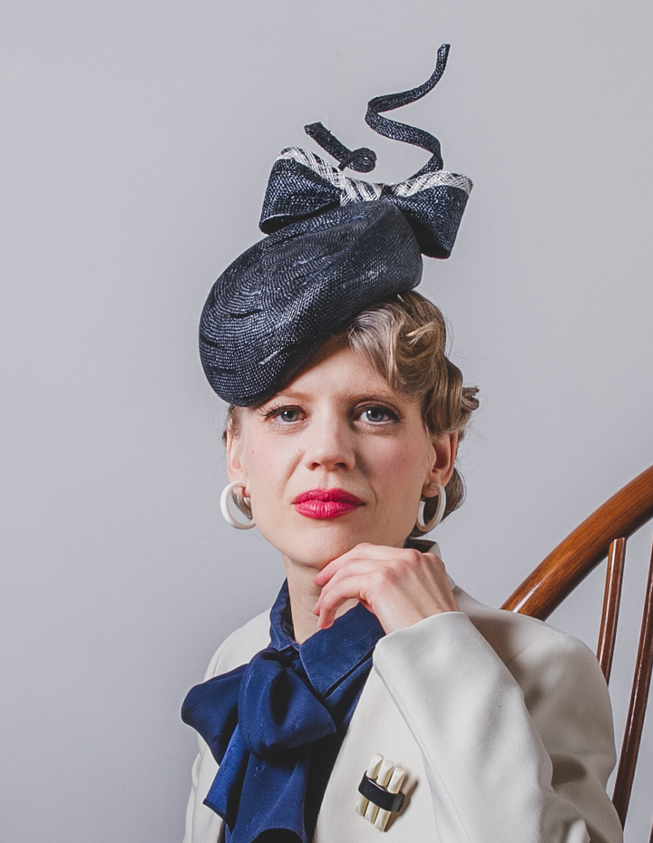 Woman wearing a decorative black hat with a large bow, white coat, and blue scarf against a gray background.