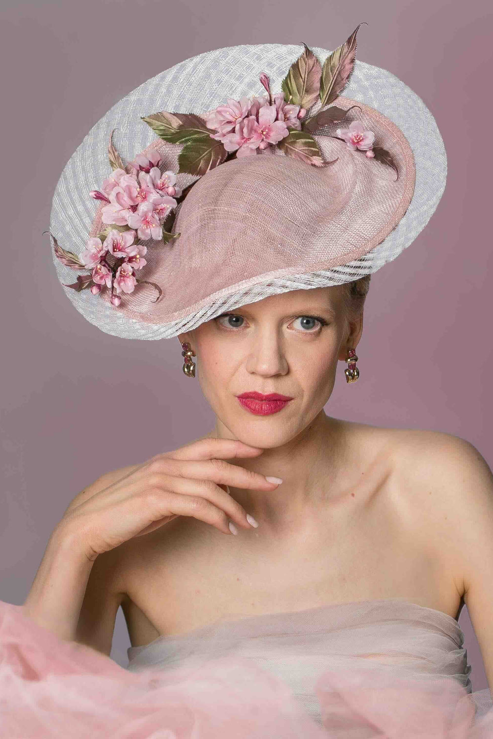 A woman wearing a white percher hat with hand-painted pink cherry blossom silk flowers and green leaves. The hat has a wire headband and a comb.
