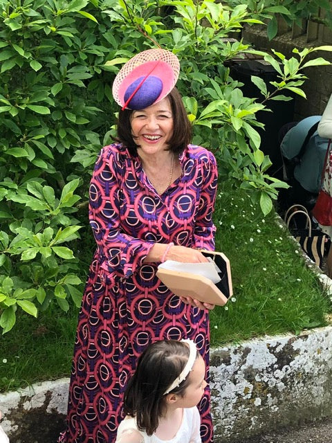 Woman in a patterned dress and colorful hat standing outdoors with greenery in the background